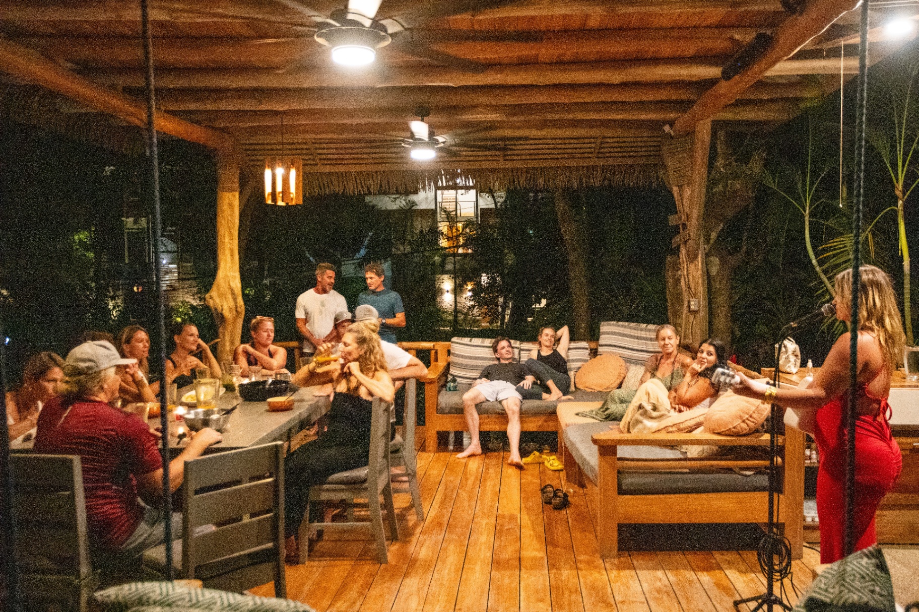 Group of guests gathered around long tables and couches in the rancho at night, sharing conversation under warm lighting.