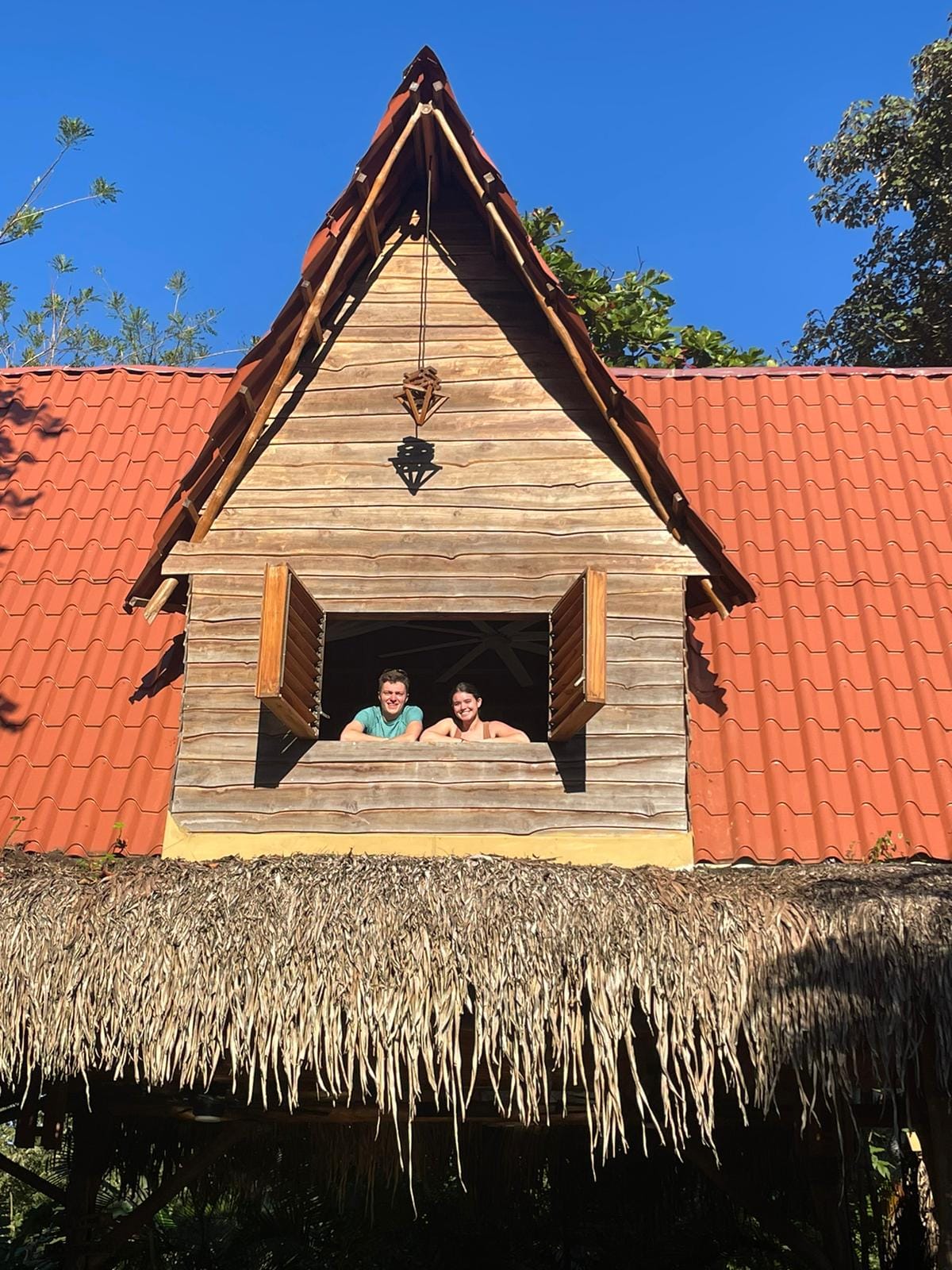 Couple smiling from the A-frame treehouse loft window against a blue sky.