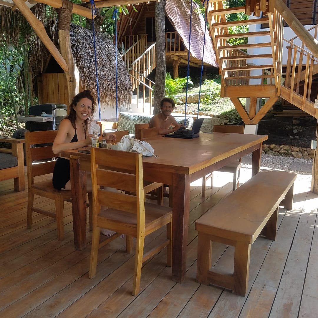 Guests at the long teak table beneath the treehouse, one on a laptop.