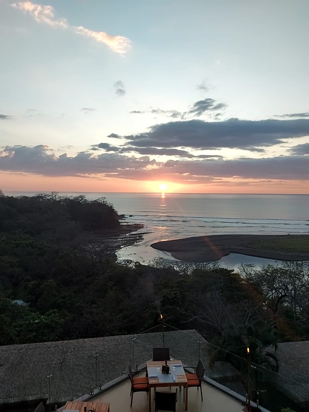 Sunset view from La Garta Lodge over the Nosara river mouth meeting the Pacific, thatched-roof terrace in the foreground.