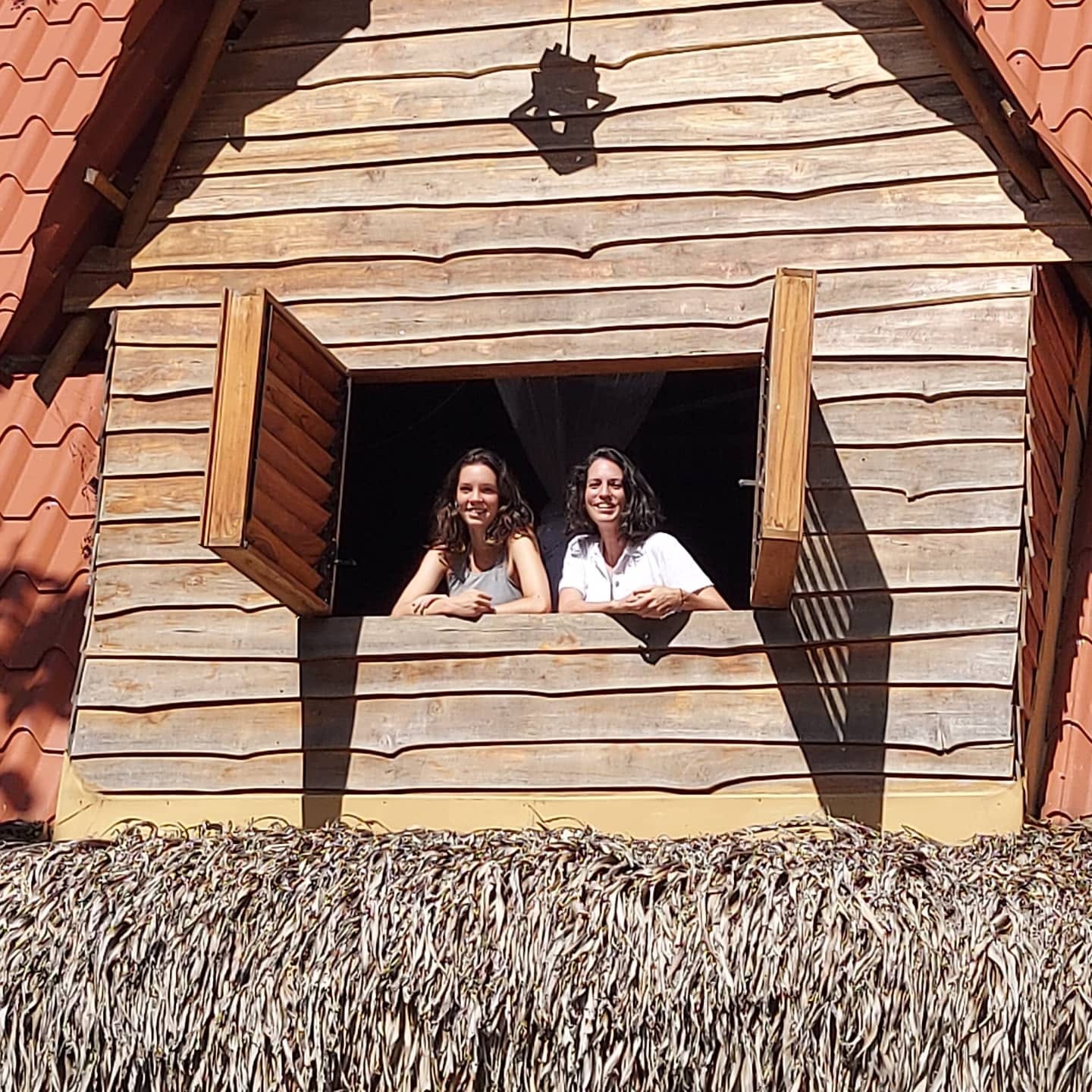 Two friends leaning out the treehouse loft window over the thatched roof.