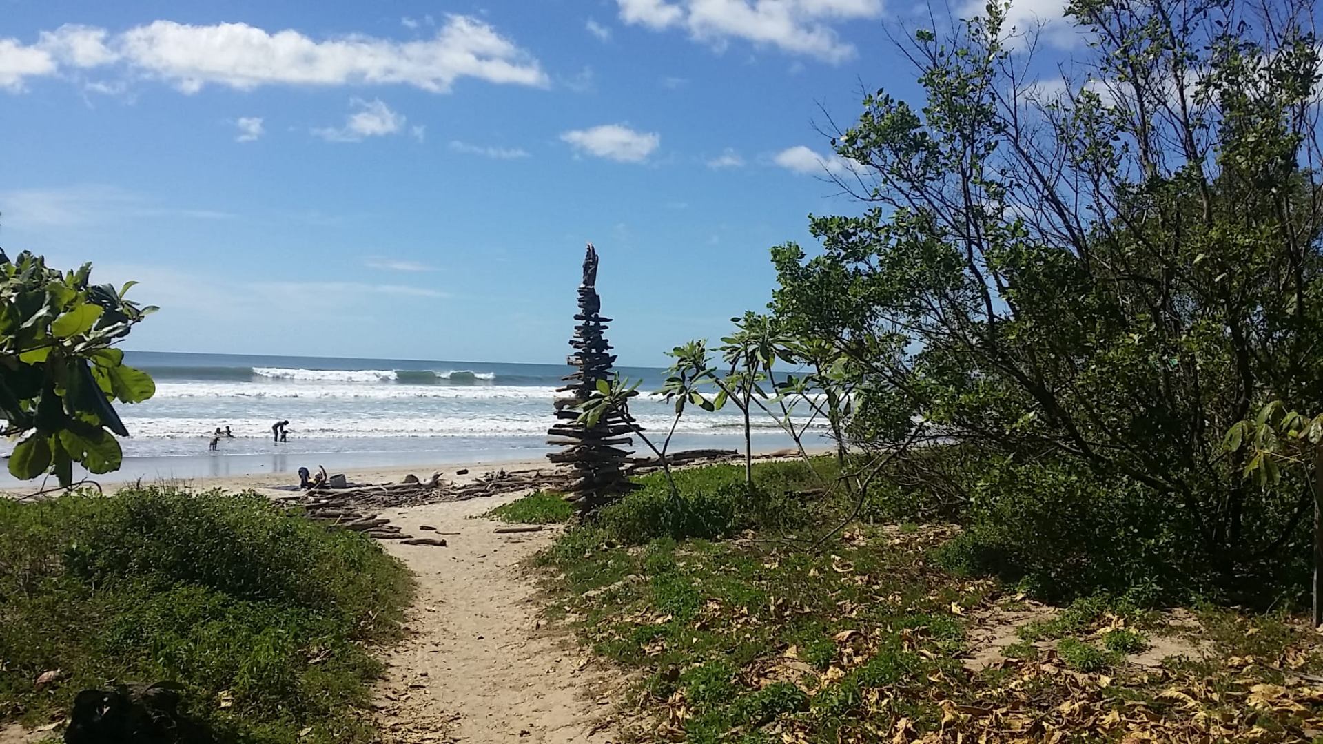 Sandy path opening onto Playa Pelada past a tall driftwood cairn, with clean surf breaking beyond.