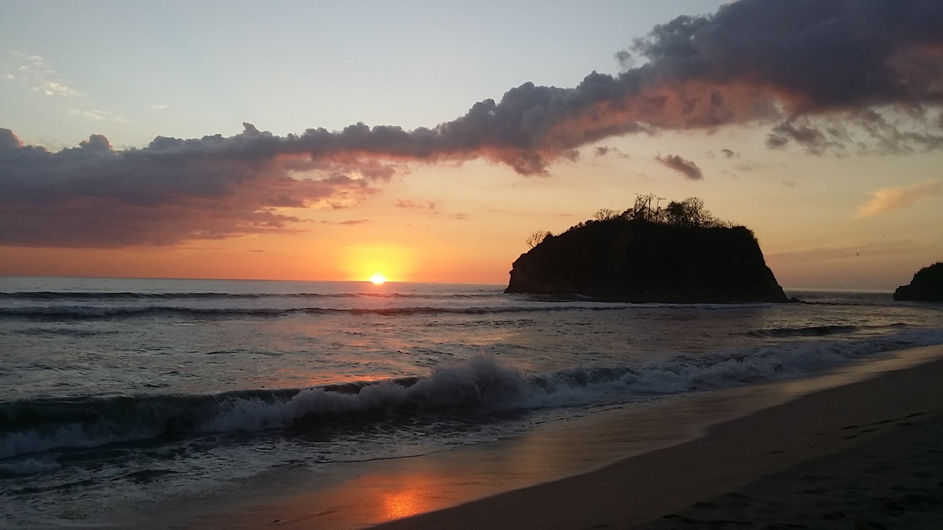 Sunset over Playa Pelada with the iconic offshore island silhouetted against a pink and gold sky.