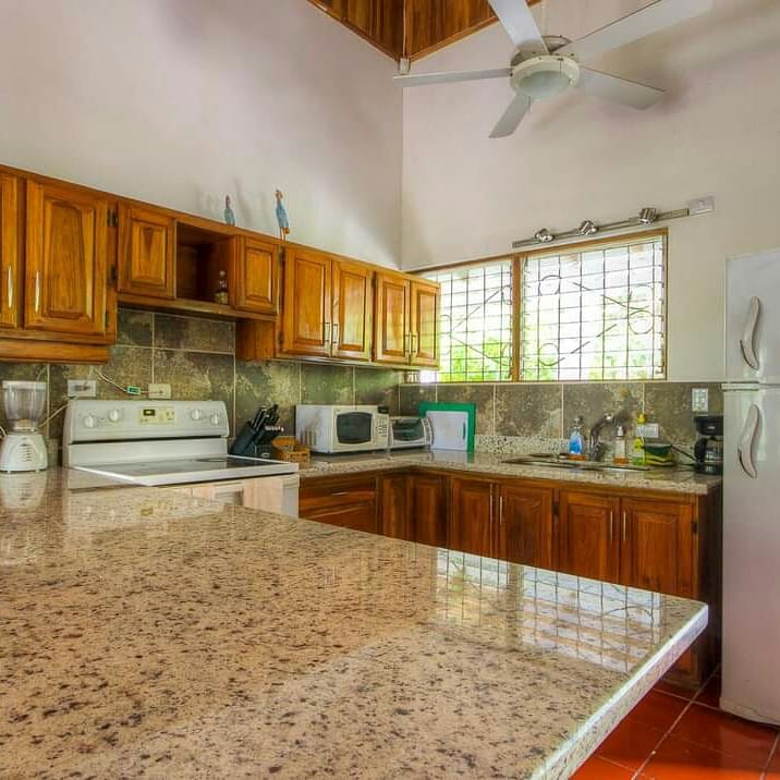 Casa Brisa kitchen before — dated wood cabinets, tile backsplash, white appliances, small barred window, terracotta floor.
