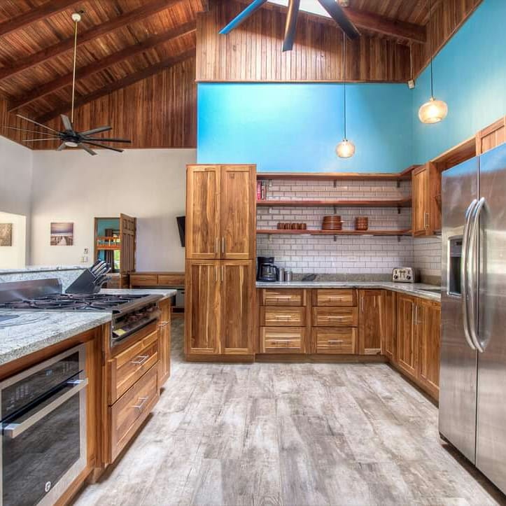Casa Brisa kitchen after — teak cabinetry, granite counters, subway-tile backsplash, teal accent wall, exposed teak-plank cathedral ceiling.
